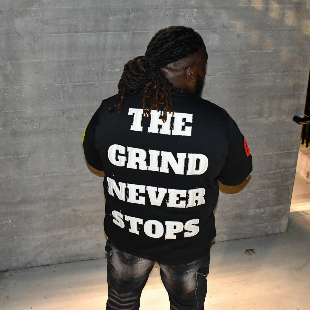 Person wearing a black t-shirt with 'THE GRIND NEVER STOPS' text on the back against a gray wall.
